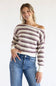 A woman modeling a chunky knit off-the-shoulder sweater with horizontal stripes in cream and dark plum, paired with blue jeans against a plain white studio background.