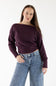 A woman modeling a dark plum purple off-the-shoulder knit sweater paired with blue jeans with a small white star pattern, against a plain white studio background.