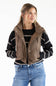 A woman wearing a brown sleeveless puffer vest over a black and white striped knit sweater, paired with blue jeans, against a plain white background.