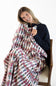 A smiling woman sits on a woven chair wrapped in a soft blanket featuring a repeating nutcracker soldier pattern on a white background.