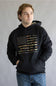 A man wearing a black pullover hoodie with a distressed American flag graphic made of camouflage-patterned stripes and the text 'SIMPLY SOUTHERN OUTFITTERS' printed below it. He is also wearing blue jeans, standing against a neutral gray studio background.