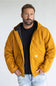 A man wearing a mustard yellow hooded work jacket over a black t-shirt and light blue jeans, standing against a light gray studio background.