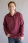 A smiling young man wearing a maroon long-sleeve quarter-zip pullover with a small black bird logo on the left chest, paired with blue jeans, standing against a neutral gray studio background.