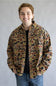 A smiling young man wearing a camouflage print jacket over a black t-shirt and blue jeans, standing against a plain gray studio background.