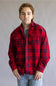 A young man wearing a red and black plaid flannel button-up shirt over a gray t-shirt and blue jeans, standing against a neutral gray studio background.