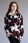 A woman with red hair wearing a brown and white cow-print fleece quarter-zip pullover, paired with black pants, standing against a neutral gray studio background.