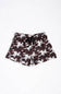 A pair of youth girls shorts featuring a dark brown and white cow pattern, with a black drawstring at the elastic waistband, laid flat against a white background.