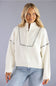 A woman wearing a cream ribbed knit half-zip sweater with black dash-like trim details, paired with light blue jeans, against a solid gray studio background.