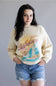Woman wearing a beige sweatshirt with a graphic design on a plain background