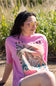 Woman wearing a pink t-shirt with a palm tree design, sitting in a natural setting.