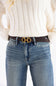 A close-up of a person wearing a dark brown leather belt with a gold-tone double-H buckle, paired with blue jeans and a cream-colored ribbed knit sweater.