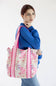 A woman wearing a blue sweatshirt and jeans carries a large tote bag with a pink and white floral pattern and pink straps, set against a plain white background.
