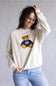 A woman wearing a cream-colored crewneck sweatshirt with a teddy bear graphic wearing a navy blue jersey with the number 98, paired with light blue jeans, against a neutral gray studio background.