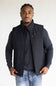 A man wearing a black zip-up insulated vest over a dark gray long-sleeve shirt and blue jeans, standing against a plain light background.