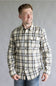 Front view of a smiling male model with hands in his pockets wearing the Split Flannel Shirt for Men in Grey Ecru, featuring a beige, gray, and brown plaid pattern, long sleeves, and dual flap chest button pockets, paired with medium-wash jeans against a neutral gray studio background.