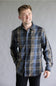 A smiling male model posing against a neutral gray studio background, wearing the Split Flannel Shirt for Men in Moka featuring a blue, dark gray, and tan plaid pattern with long sleeves and dual buttoned chest pockets.