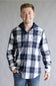 Model wearing the Split Men's long-sleeve hooded flannel shirt in a navy, ecru white, and tan plaid pattern, featuring two chest flap pockets and a solid navy attached hood with drawstrings against a gray studio background.