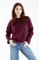 A woman with red hair wearing a loose-fitting, crewneck knit sweater in deep burgundy and blue jeans, standing against a plain white studio background.