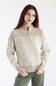 A woman with long red hair wearing a beige and white horizontally striped quarter-zip sweater and olive green pants, standing against a plain white studio background.