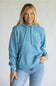 A woman wearing a light blue pullover hoodie with a small white embroidered logo on the left chest, paired with light-wash blue jeans, standing against a plain light gray background.