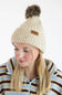A woman wearing a cream ribbed knit pom-pom beanie with a small brown brand patch and a blue, brown, and cream striped fleece zip-up sweater, photographed against a clean white background.