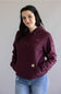 A woman with long dark hair smiling, wearing a deep cherrywood Carhartt hoodie with a front kangaroo pocket, a small chest logo, and the Carhartt logo printed along the left sleeve, paired with light-wash denim jeans.