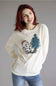 A smiling woman with red hair wearing a cream long-sleeve t-shirt with a cartoon graphic of a character next to a pine tree and the word 'Carhartt' printed below it, paired with blue jeans against a plain gray studio background.