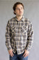 A man wearing a beige and navy plaid long-sleeve button-up shirt with two chest pockets, paired with blue jeans, standing against a neutral gray studio background.