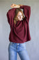 A woman wearing a maroon ribbed knit long-sleeve sweater and blue jeans, posing with her arms raised against a plain gray studio background.