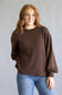 A woman with red hair wearing a brown waffle-knit long-sleeve sweater and blue jeans, standing against a neutral gray studio background.
