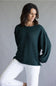 A woman wearing a dark forest green ribbed knit sweater with balloon sleeves and white pants, posing against a neutral gray studio background.