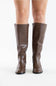 A pair of knee-high brown faux-leather boots with a low block heel, shown on a model's feet against a plain white background.