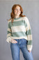 A woman with red hair wearing a fuzzy knit sweater with horizontal sage green and cream stripes, paired with blue jeans, standing against a solid gray studio background.