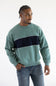 A man wearing a sage green crewneck sweatshirt with a thick navy blue horizontal stripe across the chest, paired with blue jeans, photographed against a plain white studio background.