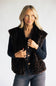 A blonde woman wearing a dark brown and black leopard-print faux fur vest over a black long-sleeve top and gray jeans, standing against a light gray studio background.