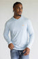 A man wearing a light blue long-sleeve hooded shirt with a small gold chest logo, paired with blue jeans, photographed against a plain light gray background.