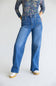 A model wearing a pair of high-waisted, wide-leg blue denim jeans with a floral long-sleeve top and white platform sneakers, standing against a plain white studio background.