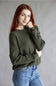 A woman with red hair wearing an olive green waffle-knit sweater and blue jeans, posing against a neutral gray studio background.