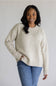 A woman wearing a cream-colored, speckled knit crewneck sweater and blue jeans, standing against a plain light gray background.