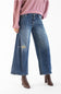 A model wearing a pair of wide-leg blue denim jeans with distressed details, paired with a dusty pink sweatshirt and brown heeled ankle boots, shown from the waist down on a white studio background.