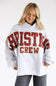 A woman wearing an oversized light gray sweatshirt with 'CHRISTINE CREW' printed across the front in red and black plaid lettering, paired with dark gray jeans, against a plain white studio background.
