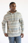 A man wearing a long-sleeve striped hoodie with a kangaroo pocket, featuring horizontal stripes in beige, brown, and gray on an off-white base, paired with blue jeans against a plain white background.