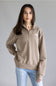 A woman wearing a taupe brown quarter-zip sweatshirt with a small white embroidered logo on the left chest, paired with light blue jeans, against a solid gray studio background.