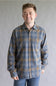 A smiling man wearing a long-sleeve plaid button-up shirt in blue and brown tones, paired with blue jeans, standing against a neutral gray studio background.