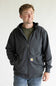 A man wearing a dark charcoal gray Carhartt zip-up hoodie over an olive green t-shirt and blue jeans, standing against a plain white background.