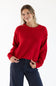 A woman wearing a red sweater with exposed seams styled with dark blue striped denim jeans standing in front of a white studio background