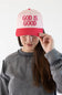 A woman wearing a pink and red trucker hat with the words 'GOD IS GOOD' embroidered in red on the front, holding the brim with her hand against a plain white background.