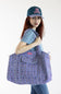 A woman wearing a blue denim t-shirt, jeans, and a navy blue baseball cap with 'DOG LOVER' embroidered in pink, carrying a large lavender and purple patterned tote bag over her shoulder, set against a plain white background.
