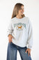 Front ¾ view of a female model wearing the Sweet Claire Embroidered Club Retriever Sweatshirt in Ash heather grey, featuring a green "Club Retriever Stay Golden" chest embroidery, crew neck, and relaxed fit paired with denim jeans.
