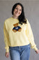 A smiling woman wearing a pale yellow crewneck sweatshirt with a teddy bear graphic wearing a navy blue jersey with the number 98, paired with blue jeans, against a neutral gray background.